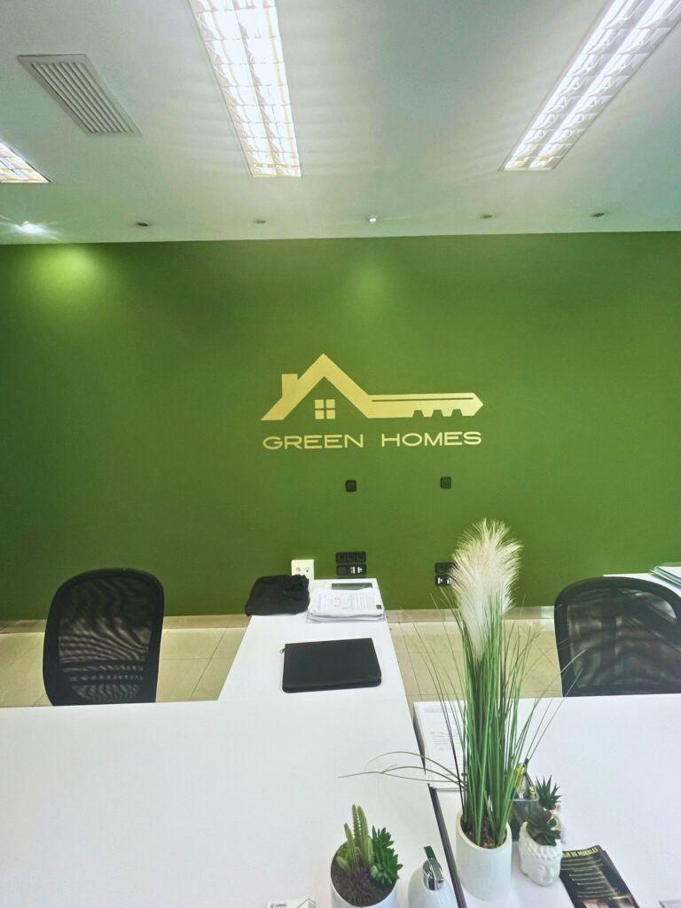 Interior de oficina Green Homes con pared verde corporativa, logotipo y mesa de atención al cliente.